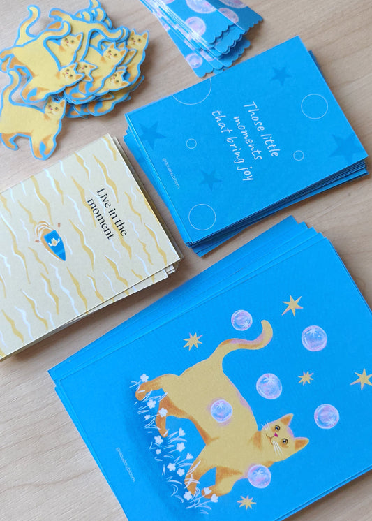 Snail mail set comes with a postcard with a yellow cat playing with bubbles on a blue background illustrations, washi sticker in a stamp shape with cats and bubble illustrations. Activity self love card and music playlist suggestion.